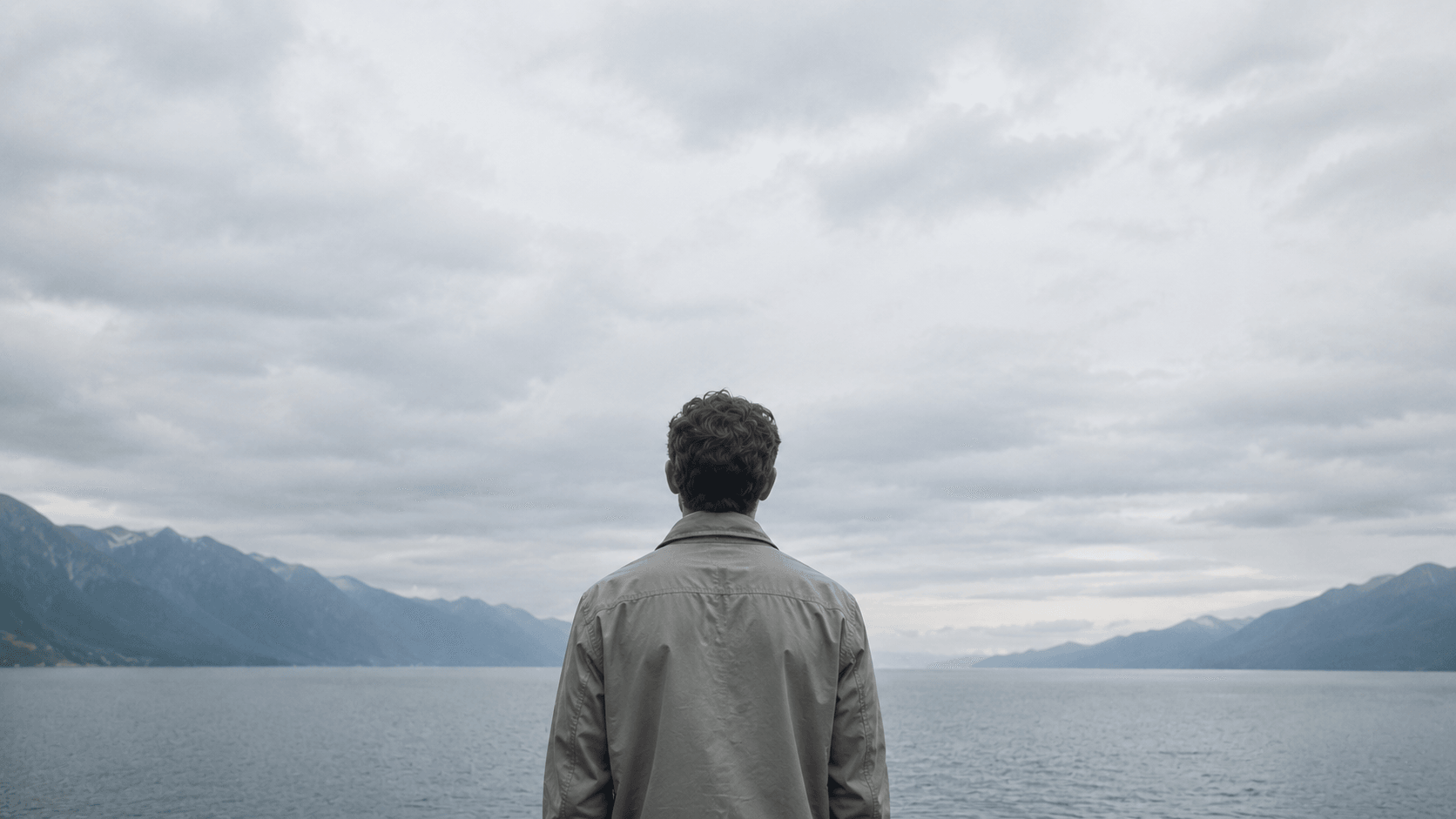 a man standing in front of the wide lake bordered by green mountains with a focus on horizon, symbolizing a path forward in mental health rooted in biology